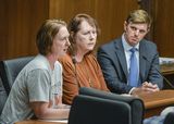 Nicole Smith-Holt sits at the testifiers' table and speaks to someone out of view. Cindy Scherer Boyd and Representative Howard sit next to her. Representative Howard watches Nicole speak.